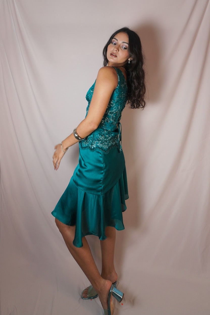 A woman modeling a teal green sleeveless dress with a lace-embellished bodice and a ruffled skirt, posing against a draped white fabric backdrop.