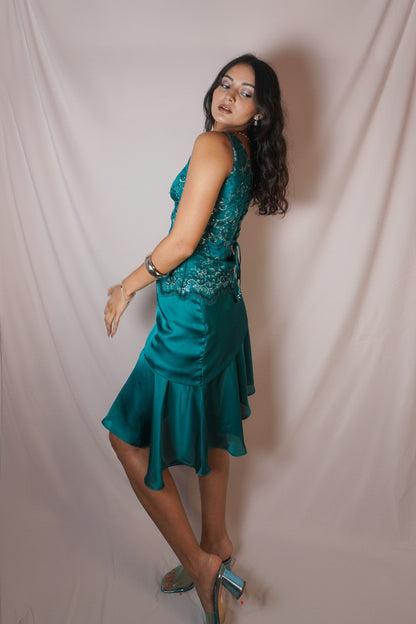 A woman modeling a teal green sleeveless dress with a lace-embellished bodice and a ruffled skirt, posing against a draped white fabric backdrop.
