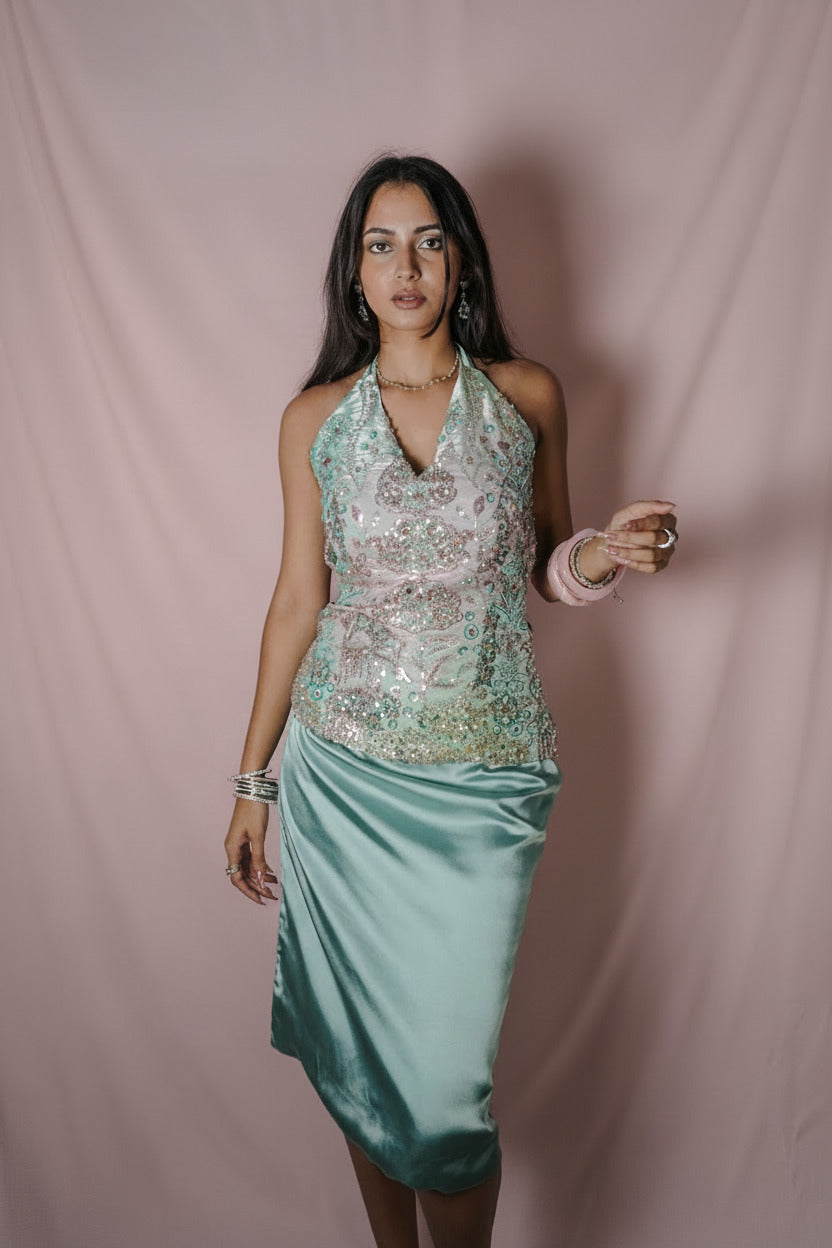 A woman modeling a sleeveless halter-neck dress with a heavily embellished light pink and green sequined bodice and a satin teal skirt, standing against a draped gray fabric backdrop.
