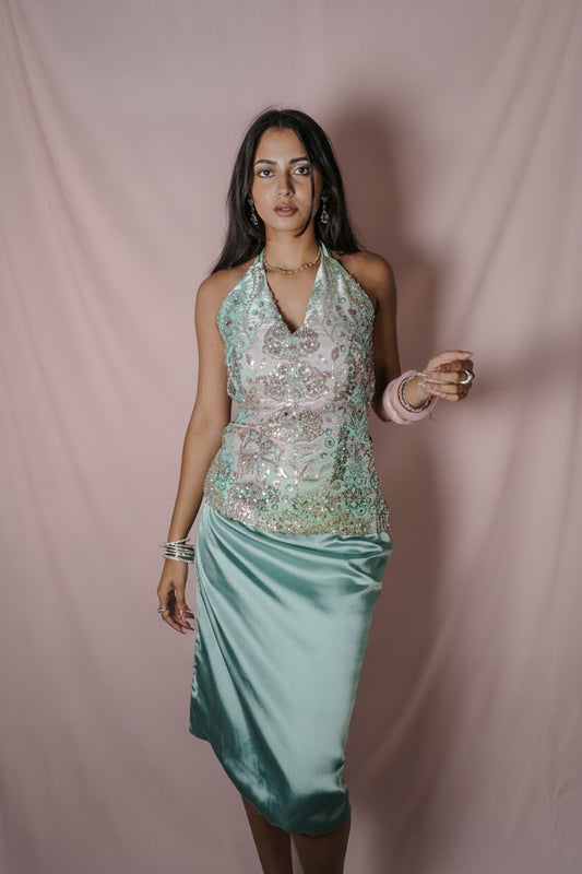 A woman modeling a sleeveless halter-neck dress with a heavily embellished light pink and green sequined bodice and a satin teal skirt, standing against a draped gray fabric backdrop.