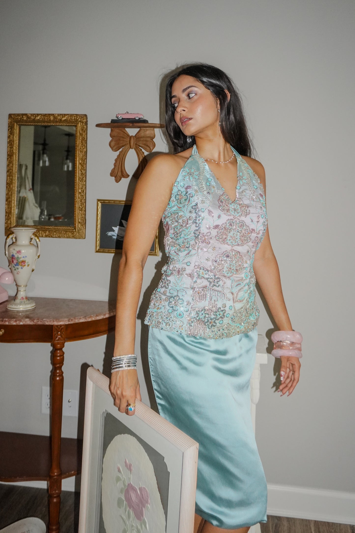 A woman models a sleeveless halter top with a light blue and pink floral lace overlay paired with a matching light blue satin midi skirt, standing in a room with vintage furniture.