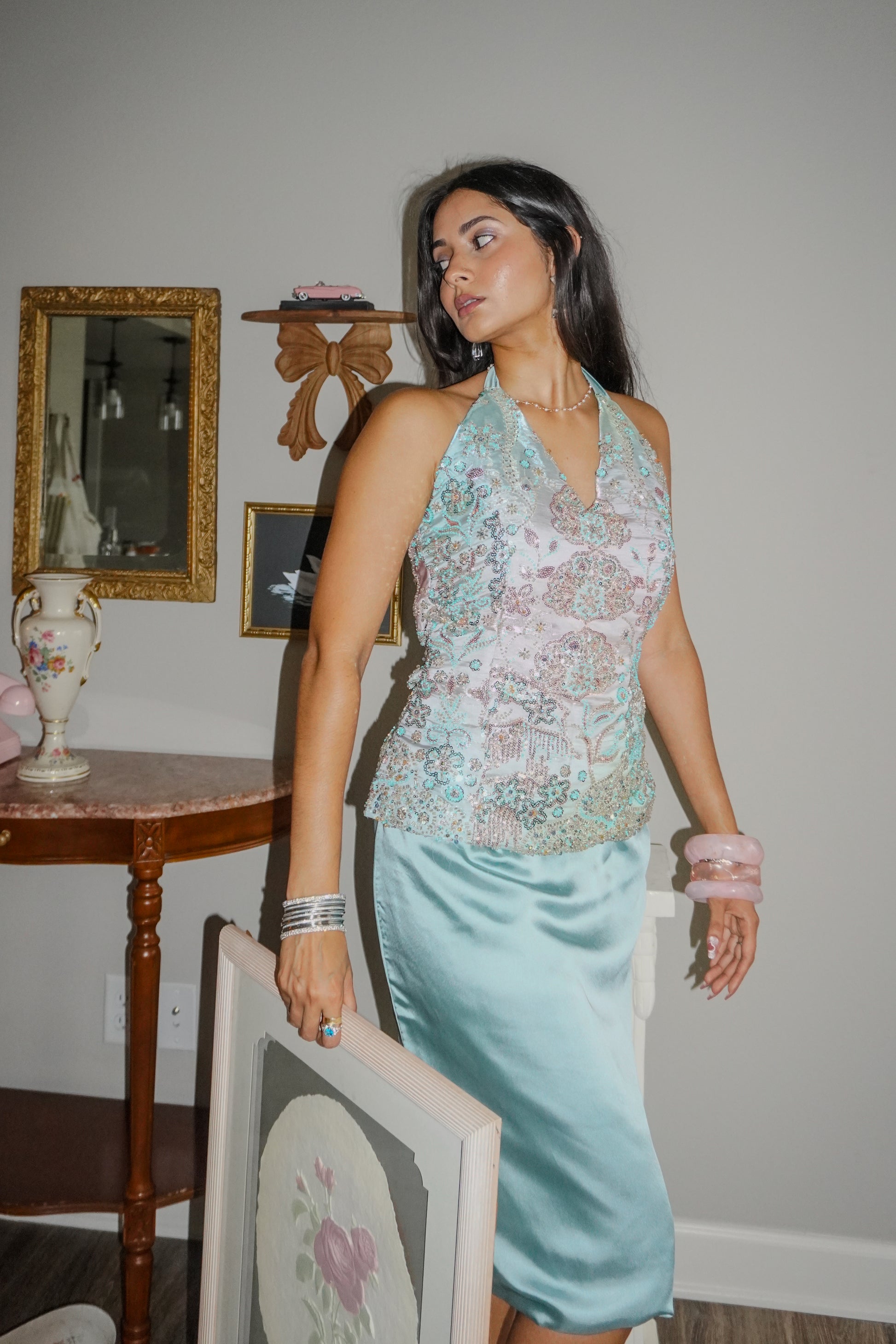 A woman models a sleeveless halter top with a light blue and pink floral lace overlay paired with a matching light blue satin midi skirt, standing in a room with vintage furniture.